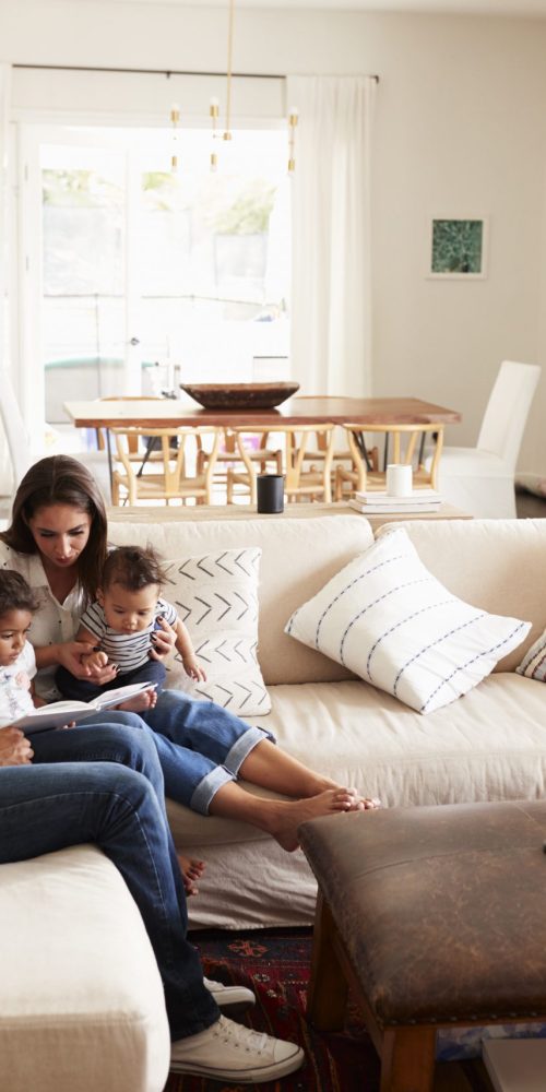 Young Hispanic family sitting on sofa reading a book together in the living room, seen from doorway