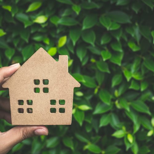 Hand holding wooden house on green leaves background with copy space. Home ecology concept.